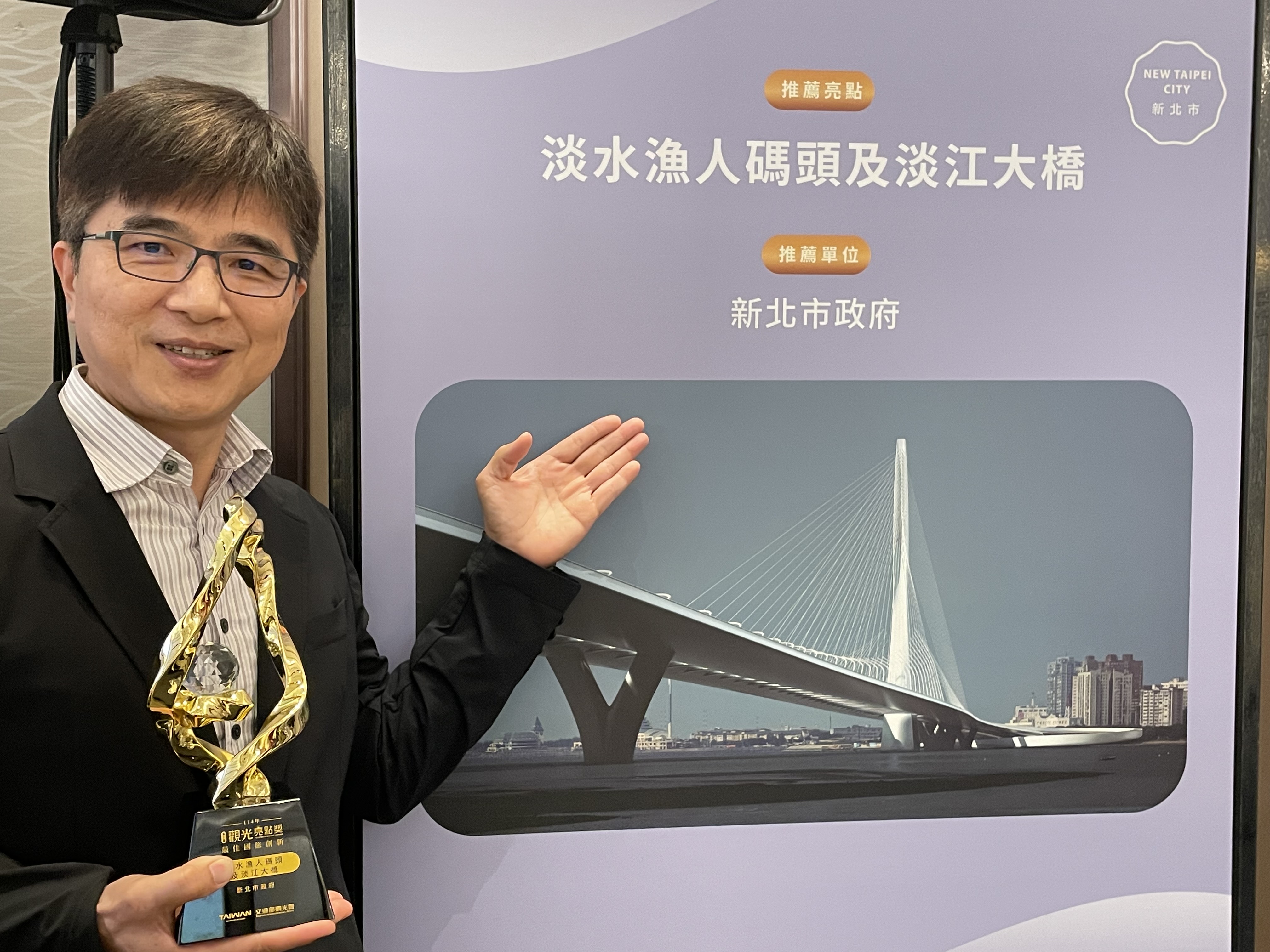  新北市觀光旅遊局莊榮哲副局長持「最佳國旅創新獎」獎盃，於淡江大橋主題背板前合影。