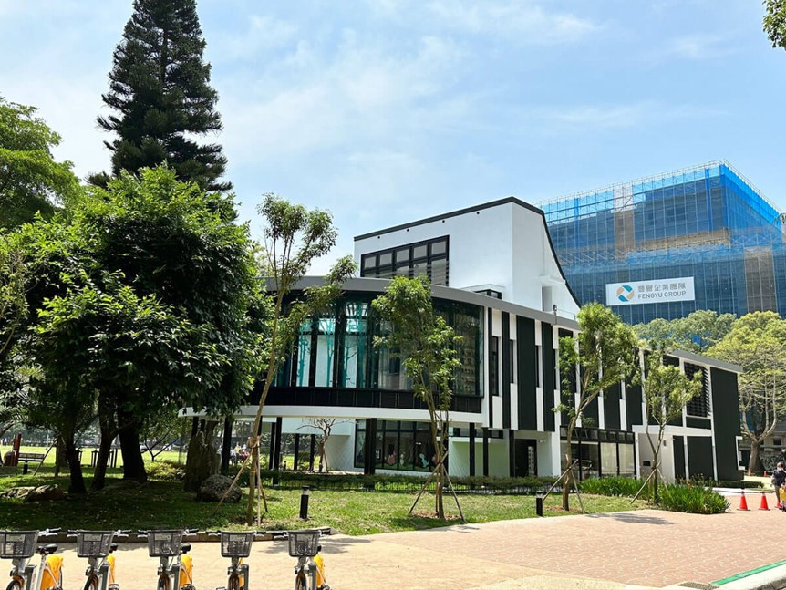  林口區李科永紀念圖書館外觀為沉穩的黑色線條及半圓型玻璃走廊矗立於綠意盎然的公園環境