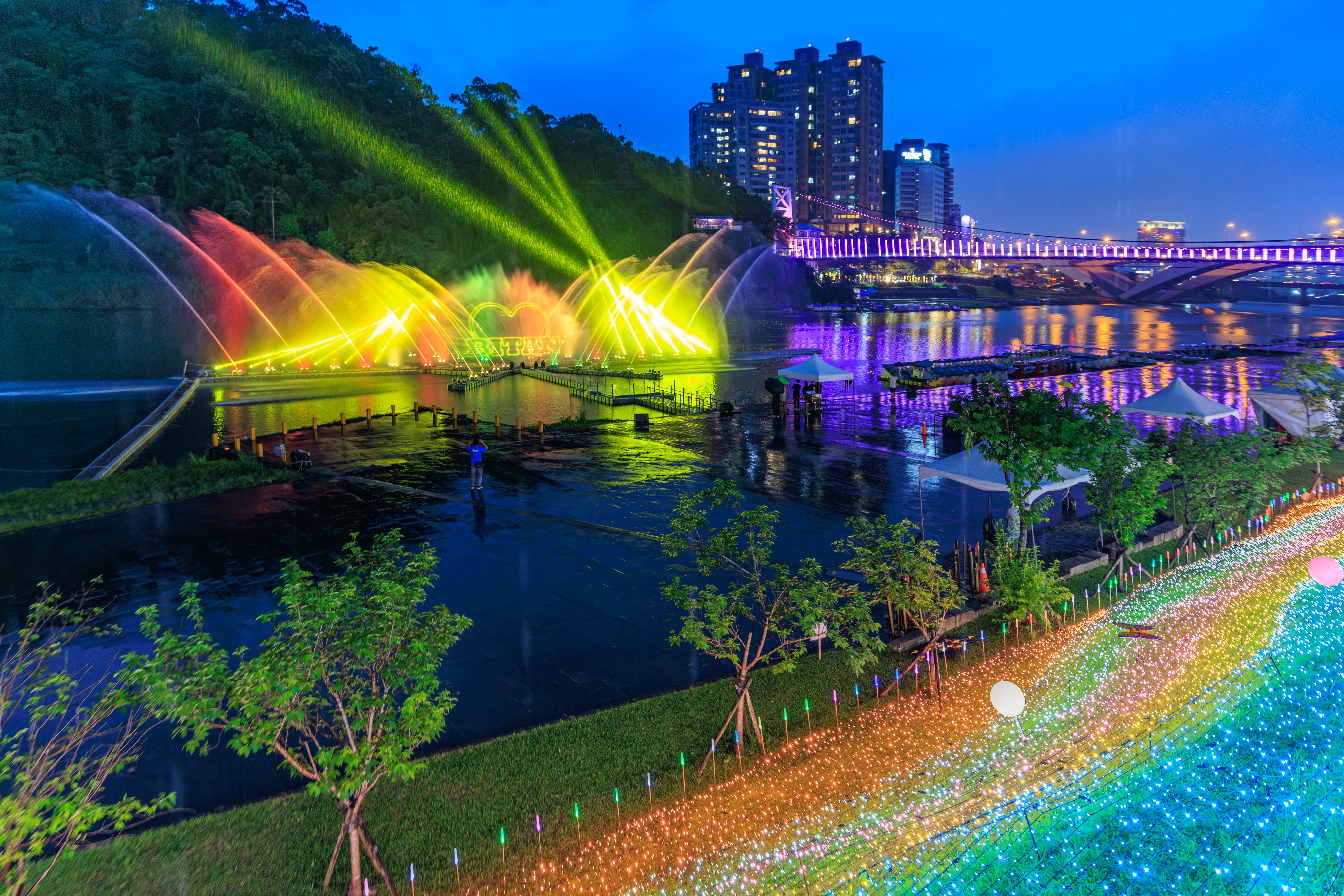  夜幕降臨，碧潭水域化身流光舞台，七彩水柱隨音樂律動綻放，與吊橋燈飾及岸邊光廊交織成一幅夢幻夜景，揭開2026碧潭水舞季序幕。(由攝影師Justin Cheng拍攝)