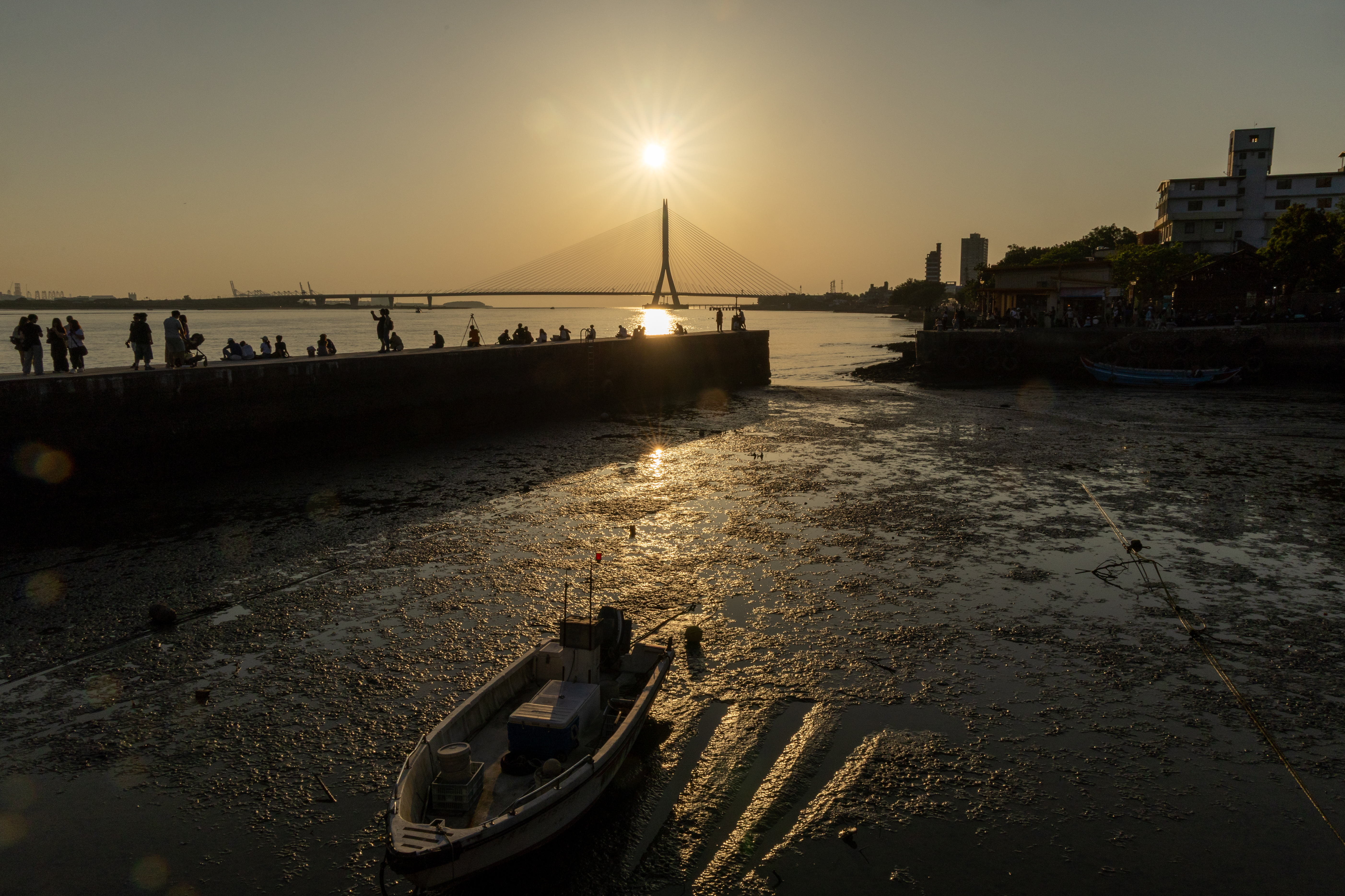  夕陽停泊於淡江大橋頂端，金黃餘暉灑落小船。淡水暮色下，現代建設與傳統日常在此交融，堤岸上的人群也化作了一道道迷人的剪影。