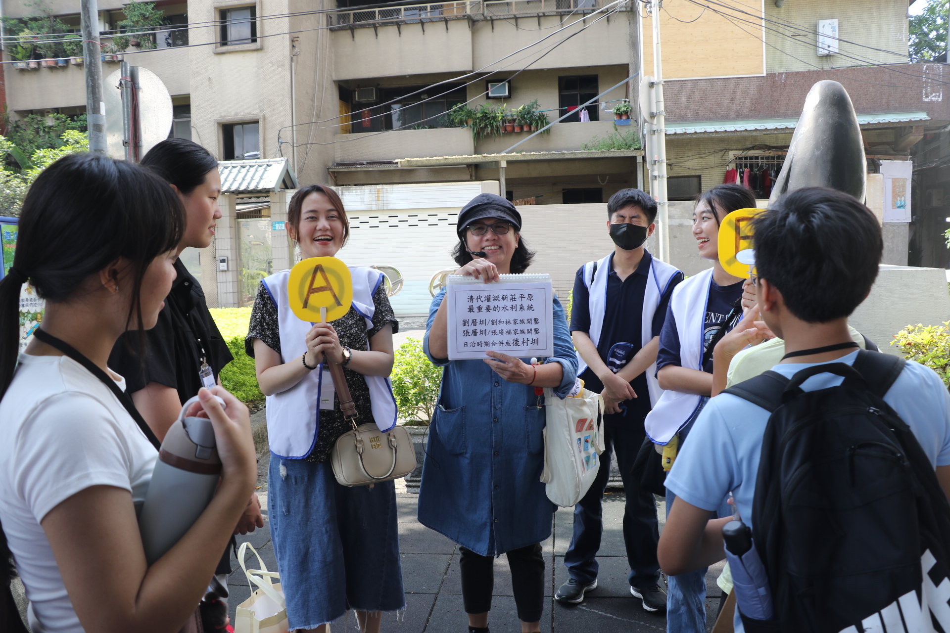  新莊社區大學講師王惠靜（中）為兒少解說先人開墾新莊平原建設的劉厝圳、張厝圳，並要兒少搶答。左3為綽號「大飽」的新莊少年福利中心主任許巽絜。