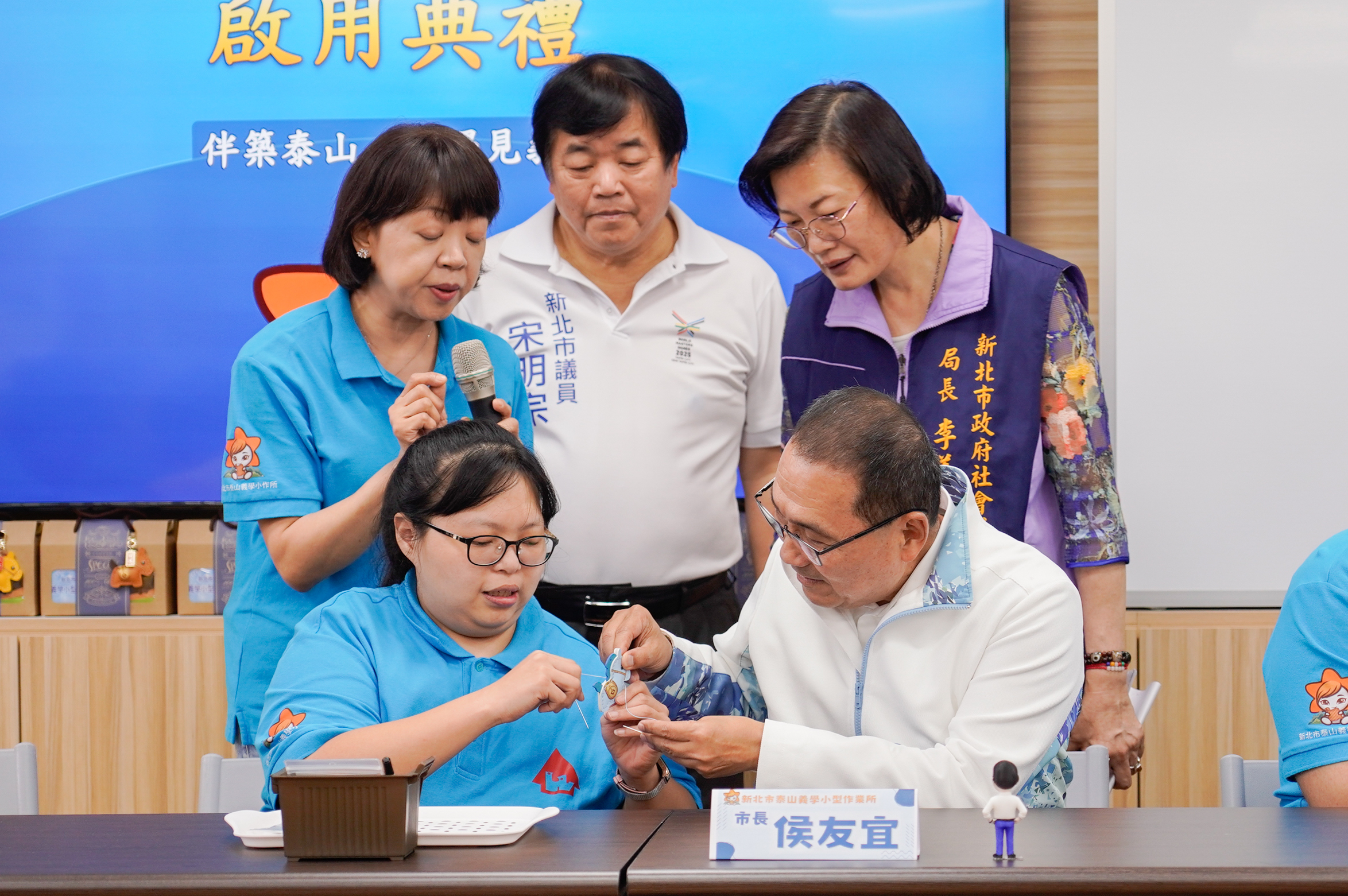侯友宜與星兒體驗皮革文創手作，由學員小玲(前排左)擔任小老師，透過穿針引線、放進包裝袋等互動，一起完成象徵平安、幸福的「好運馬」。圖後排右起為社會局長李美珍、議員宋明宗。